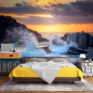 May include: A bedroom with a bed and a headboard against a wall mural depicting a rocky seascape with crashing waves at sunset. The mural features a dramatic sky with orange, yellow, and purple clouds. The bed has a yellow and white comforter and a gray pillow. A small table with a lamp and a basket is next to the bed.