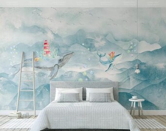 May include: A watercolor mural featuring two blue whales swimming in a blue and white seascape. The mural is painted on a wall behind a gray bed with white bedding. The mural also features a red and white lighthouse.