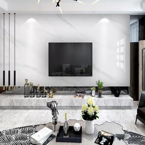 Puede incluir: Un salón moderno con un televisor de pantalla plana grande montado en una pared de mármol blanco. La habitación cuenta con una consola con tapa de mármol con objetos decorativos, una bandeja negra con una botella y un objeto geométrico, y un jarrón blanco con flores amarillas.