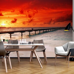 May include: A dining room with a wooden table and chairs set against a backdrop of a sunset over a tropical ocean with a pier extending into the water.