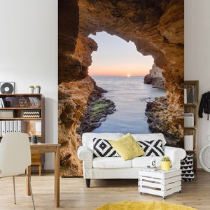 May include: A white couch with black and white patterned pillows sits in front of a wall mural depicting a rocky cave opening to a view of the ocean at sunset.