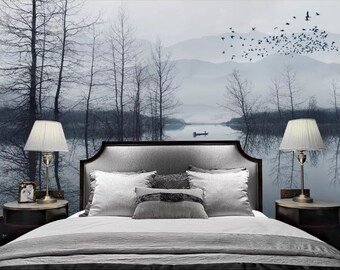May include: A bedroom with a gray upholstered bed with white pillows and a gray blanket. The bed is in front of a wall with a scenic wallpaper depicting a misty lake with trees and birds flying overhead. Two lamps with white lampshades are on nightstands on either side of the bed.