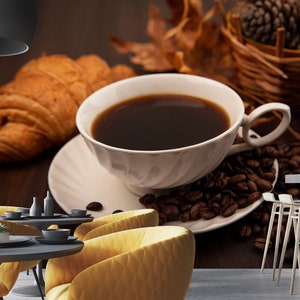 Puede incluir: Una taza de café de cerámica blanca llena de café oscuro se encuentra sobre un platillo con granos de café. Un croissant está en el fondo.