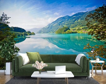 May include: A green sofa with white throw cushions sits in front of a scenic wall mural depicting a lake surrounded by lush green mountains. The lake reflects the blue sky and white clouds.