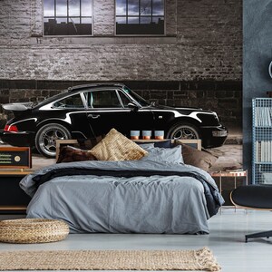 May include: A black sports car is the focal point of this bedroom wall mural. The car is parked in front of a brick wall. The bed has gray bedding and decorative pillows. A wooden radio sits on a black cabinet.