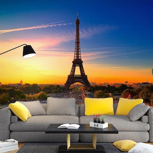 May include: A gray couch with yellow throw pillows sits in front of a wall mural depicting the Eiffel Tower in Paris, France at sunset. The mural shows the tower in the distance with a cityscape in the foreground.