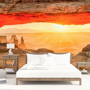 May include: A scenic sunset view of a desert landscape framed by a large red rock archway. The sun is setting behind the mountains, casting a warm glow over the scene.