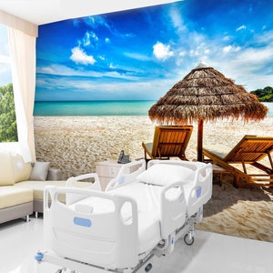 May include: A white hospital bed with a tropical beach scene mural on the wall behind it. The mural features a blue sky, white clouds, a sandy beach, turquoise water, and a thatched-roof umbrella with two lounge chairs.