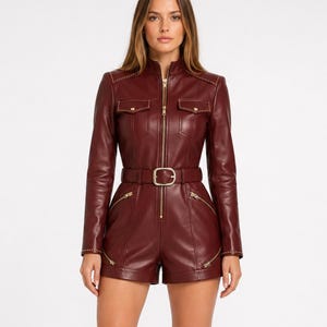 May include: A deep burgundy long-sleeve romper featuring a front zipper closure, a stand-up collar, and a matching belt with a gold-tone buckle. The garment includes two chest pockets with flaps and two angled side pockets, all accented with gold-tone studs.
