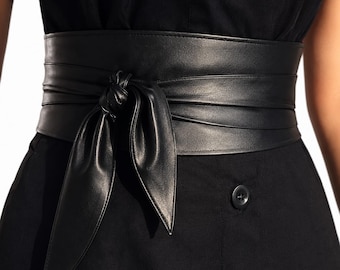 Könnte beinhalten: Ein breiter schwarzer Gürtel im Obi-Stil aus glattem Material mit Wickeldesign, der vorne mit einem Knoten gebunden wird. Der Gürtel wird über einem einfarbigen schwarzen Kleidungsstück getragen, das unterhalb der Taille einen sichtbaren Knopf aufweist.