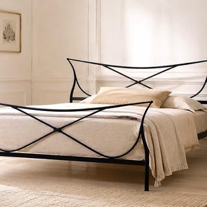 May include: Black metal bed frame with an arched headboard and footboard. The bed is made up with beige bedding and pillows. A small black side table with a lamp and a mug sits beside the bed. The room has a neutral color palette.