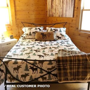 May include: A bed with a black metal frame, covered with a quilt and pillows featuring moose and pine tree patterns. A brown plaid blanket is folded at the foot of the bed. The room has wood paneling.