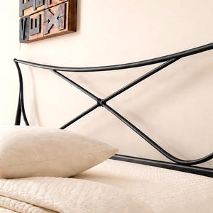 May include: Black metal bed headboard with a curved design. The headboard has an X-shaped detail in the center and is positioned above a bed with a cream-colored pillow and bedding. A decorative wooden piece hangs on the wall.