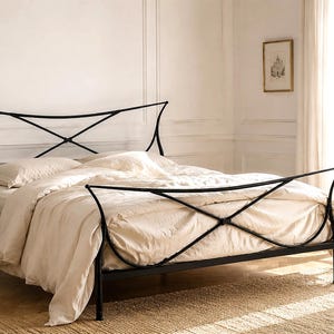 May include: A black metal bed frame with a decorative headboard and footboard. The bed is made up with cream-colored bedding. A small side table with a black lamp and a mug sits beside the bed. The room has a neutral color palette.