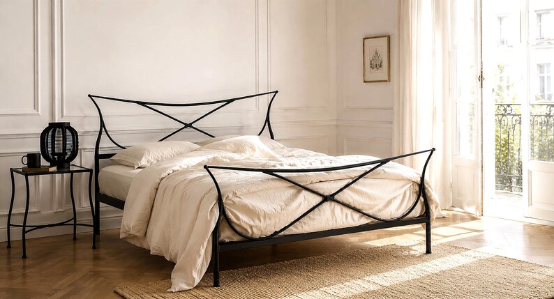 May include: A black metal bed frame with a decorative headboard and footboard. The bed is made up with cream-colored bedding. A small side table with a black lamp and a mug sits beside the bed. The room has a neutral color palette.