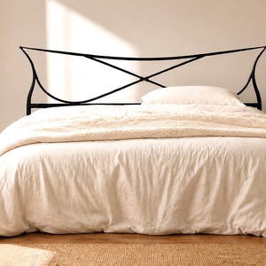 May include: A bed with a black metal headboard featuring an intersecting design. The bed is covered with a cream-colored duvet and a fluffy blanket. A small side table with a vase and decor is next to the bed.