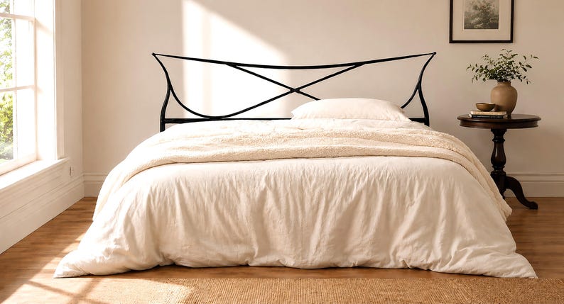 May include: A bed with a black metal headboard featuring an intersecting design. The bed is covered with a cream-colored duvet and a fluffy blanket. A small side table with a vase and decor is next to the bed.