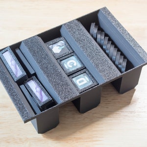 Puede incluir: Un organizador de plástico negro con compartimentos para guardar objetos pequeños. El organizador tiene una superficie texturizada e incluye una fila de pequeñas baldosas negras con letras.