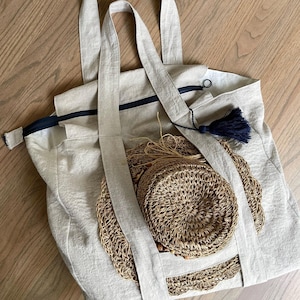 Puede incluir: Una bolsa de lona beige con cierre de cremallera y un borla negra. La bolsa está llena de dos sombreros de paja tejidos.