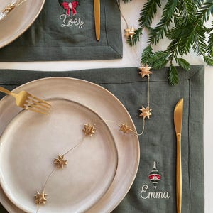 May include: Dark green linen placemats featuring embroidered holiday motifs and names. One placemat shows a wreath with the name Zoey, and the other shows an ornament with the name Emma. Each setting includes a beige plate, gold fork, and gold knife.