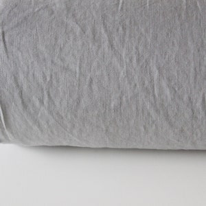 May include: A rolled-up bolt of gray linen fabric. The fabric has a textured appearance with visible weave details and a raw, unfinished edge. The color is a muted, neutral gray, suitable for various textile projects.