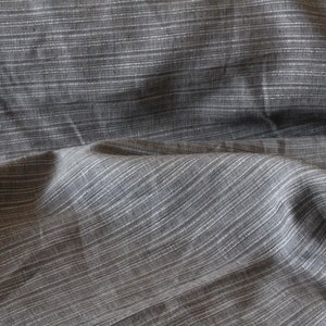 May include: Close-up of gray fabric with a subtle striped pattern. The fabric has a textured appearance with thin, horizontal white lines running across the surface. The material appears to be soft and drapes naturally, suggesting it could be used for clothing or home decor.