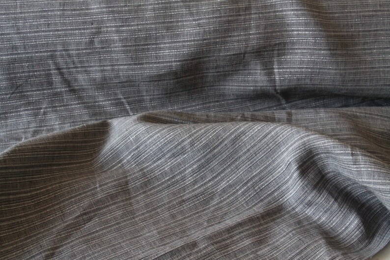 May include: Close-up of gray fabric with a subtle striped pattern. The fabric has a textured appearance with thin, horizontal white lines running across the surface. The material appears to be soft and drapes naturally, suggesting it could be used for clothing or home decor.