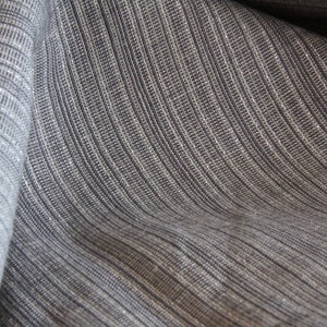 May include: Close-up of a textured fabric with a woven appearance. The fabric is a blend of gray and white threads, creating a subtle striped pattern. The material is draped, showing the texture and the way it catches the light.