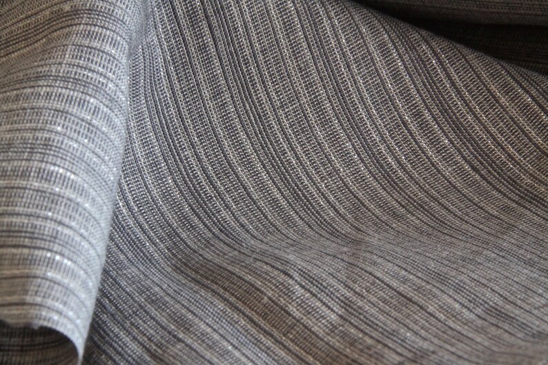 May include: Close-up of a textured fabric with a woven appearance. The fabric is a blend of gray and white threads, creating a subtle striped pattern. The material is draped, showing the texture and the way it catches the light.