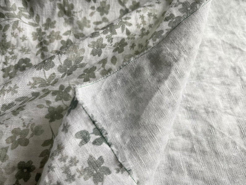 May include: Close-up of two pieces of fabric, one with a light gray background and a subtle green floral pattern, and the other a solid light gray. The fabrics are folded over each other, showing the seam where they are sewn together.