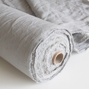 May include: A roll of light gray linen fabric. The fabric is loosely rolled, revealing the raw edges of the material. A portion of the fabric is unrolled, showing the texture and weave. The fabric is suitable for sewing and crafting projects.