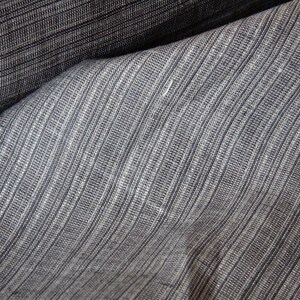 May include: Close-up of a textured fabric with a striped pattern. The fabric is a blend of gray and dark blue, with vertical lines creating a subtle ribbed effect. The material appears to be folded, showing the texture and depth of the weave.