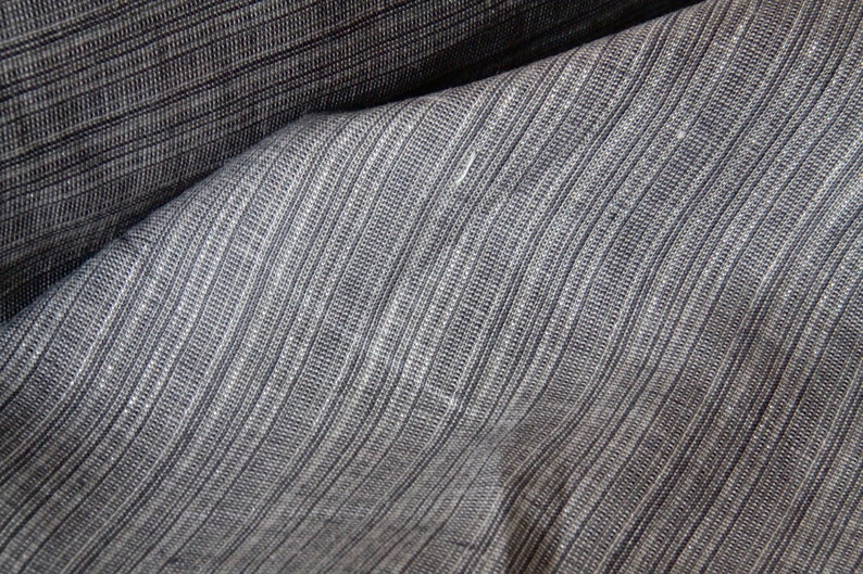 May include: Close-up of a textured fabric with a striped pattern. The fabric is a blend of gray and dark blue, with vertical lines creating a subtle ribbed effect. The material appears to be folded, showing the texture and depth of the weave.
