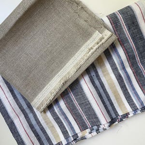 May include: Two pieces of fabric are displayed. One piece is a solid, neutral beige linen with a visible woven texture and a frayed edge. The second piece of fabric features a vertical striped pattern in shades of dark gray, blue, white, and tan.