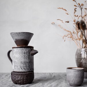 May include: A ceramic pour-over coffee maker with a matching cup and a small vase with dried plants. The coffee maker is gray and brown with a unique textured design. The cup and vase have a similar color scheme.