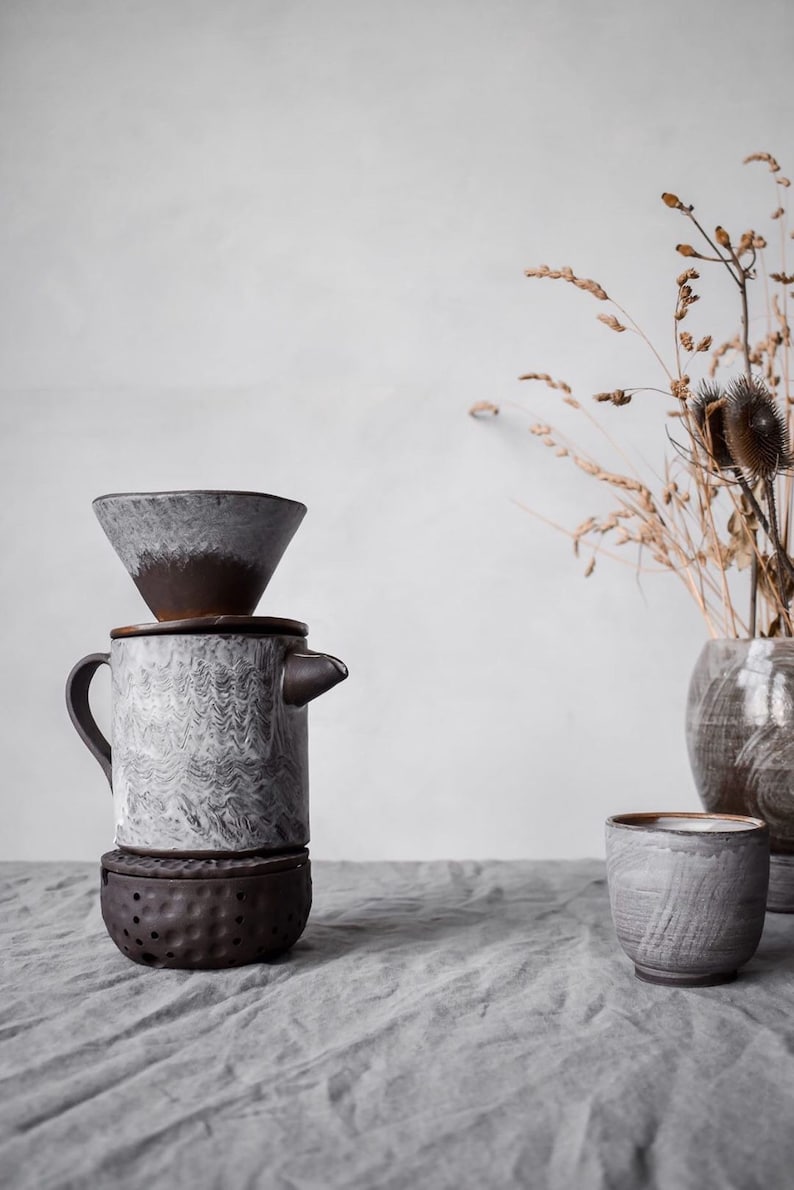 May include: A ceramic pour-over coffee maker with a matching cup and a small vase with dried plants. The coffee maker is gray and brown with a unique textured design. The cup and vase have a similar color scheme.