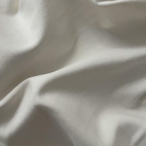 May include: A close-up view of off-white fabric featuring a distinct diagonal twill weave pattern. The material is draped in soft, flowing folds, creating natural shadows and highlights that emphasize the texture and structure of the textile.