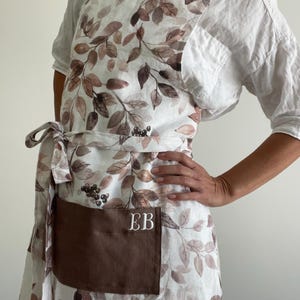 May include: An apron featuring a white base with a brown and taupe leaf and berry pattern. The apron includes a solid brown rectangular pocket on the front with the white embroidered letters EB. It has a tie waist and a cross-back strap design.