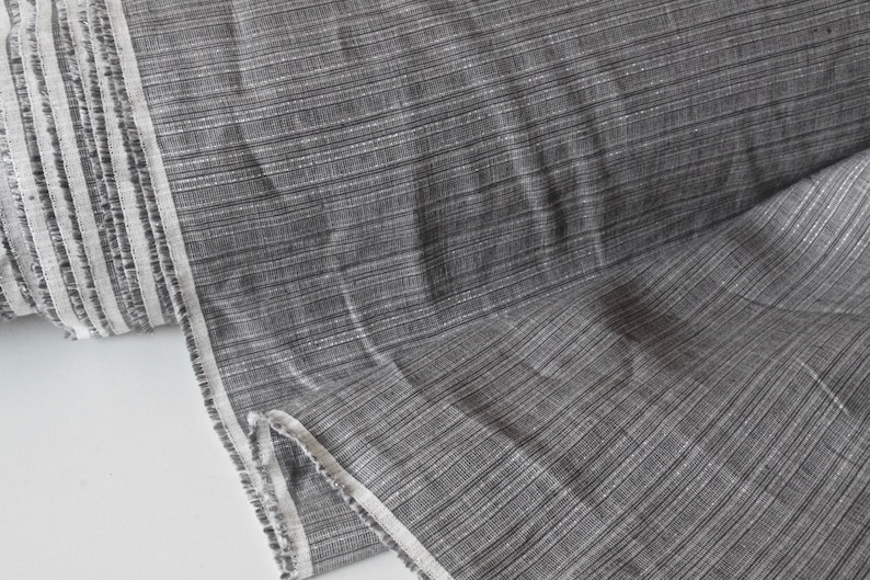May include: A roll of gray fabric with a subtle striped pattern. The fabric has a textured appearance with a light sheen, and a white border runs along the edge. The material appears to be a woven textile, suitable for various sewing projects.