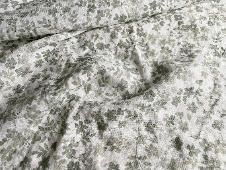 May include: White linen fabric with a gray floral pattern. The fabric is wrinkled and has a soft, textured feel.