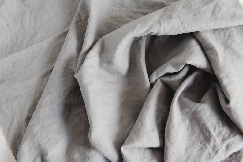 May include: A close-up shot of a gray fabric with a soft, wrinkled texture. The fabric is draped and folded, creating shadows and highlights that emphasize its texture. The overall appearance is of a light, flowing material.