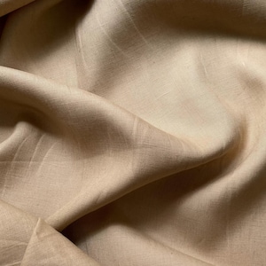 May include: A close-up of a beige linen fabric with a smooth, textured surface. The fabric is folded and draped, showcasing its soft and natural texture.