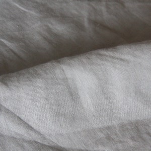May include: Close-up of a light gray linen fabric with a textured weave. The fabric is folded, creating shadows and highlighting the natural variations in the material. The overall appearance is soft and slightly wrinkled.