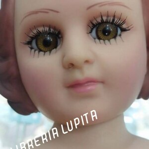 May include: A close-up of a doll's face with brown hair, brown eyes, and a pink mouth. The doll is wearing a white dress and has a light pink complexion. The text "LIBRERIA LUPITA" is visible in the lower right corner of the image.