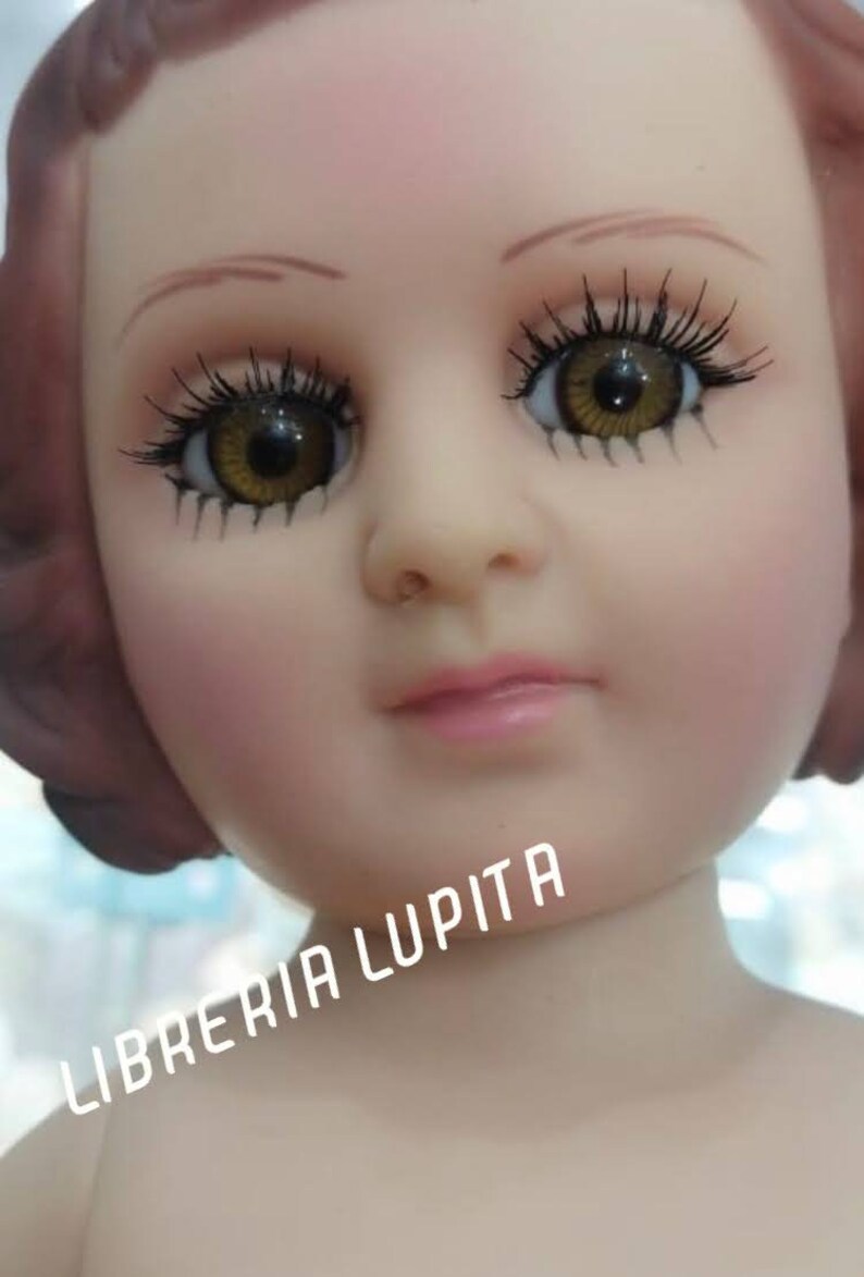 May include: A close-up of a doll's face with brown hair, brown eyes, and a pink mouth. The doll is wearing a white dress and has a light pink complexion. The text "LIBRERIA LUPITA" is visible in the lower right corner of the image.