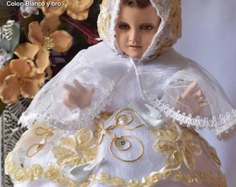 May include: White and gold baby Jesus doll wearing a white gown with gold lace trim and a white cape with gold trim. The doll is wearing a white crown with gold trim. The doll is sitting on a white surface. The text "Niño Dios con ROPÓN DE GALA" is visible at the top of the image. The text "Novedades Modelo: 5 Talla: 12 a 45 Color: Blanco y oro" is visible at the bottom of the image.