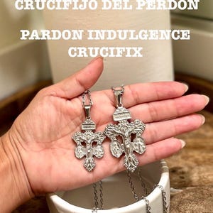 Puede incluir: Dos colgantes de crucifijo plateados sobre cadenas finas. Los colgantes representan a Jesús en la cruz, con otras figuras religiosas. La imagen incluye el texto "CRUCIFIJO DEL PERDÓN, PARDON INDULGENCE, CRUCIFIX".