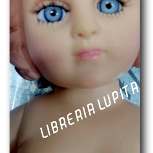 May include: A close-up of a doll's face with blue eyes and a light pink mouth. The doll is wearing a white dress and has the text "LIBRERIA LUPITA" printed on its chest.