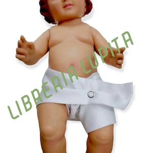 May include: A small, white-clad baby doll with a red-brown wig and blue eyes. The doll is lying on its back with its arms outstretched. The doll is wearing a white diaper with a button closure. The doll is a religious figure, likely representing the baby Jesus.