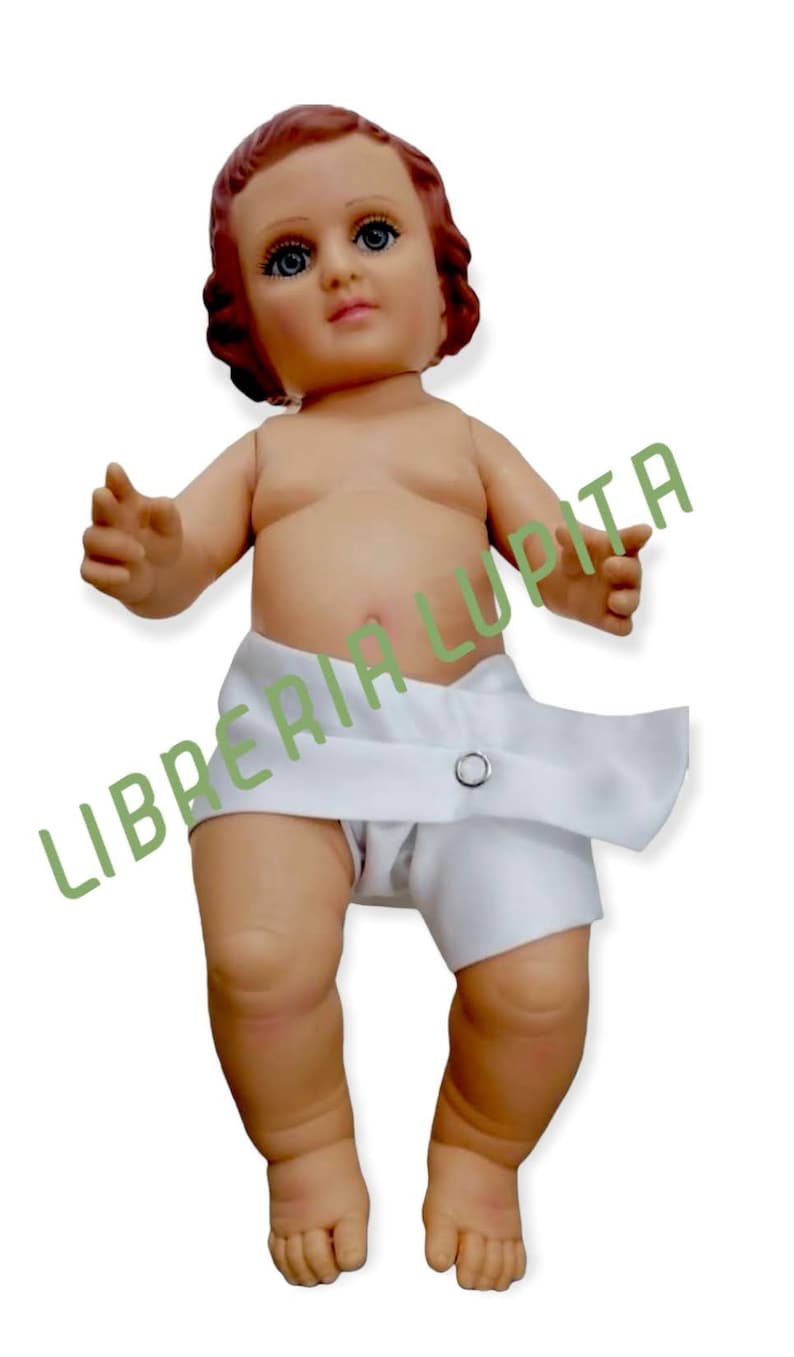 May include: A small, white-clad baby doll with a red-brown wig and blue eyes. The doll is lying on its back with its arms outstretched. The doll is wearing a white diaper with a button closure. The doll is a religious figure, likely representing the baby Jesus.